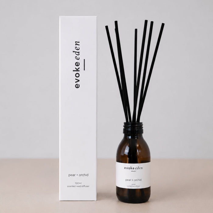 A reed diffuser set with an amber glass bottle, black reeds, a white product box, and a white label, all branded 'evokeeden' and labeled 'pear + orchid', photographed on a light beige surface against a neutral gray background.